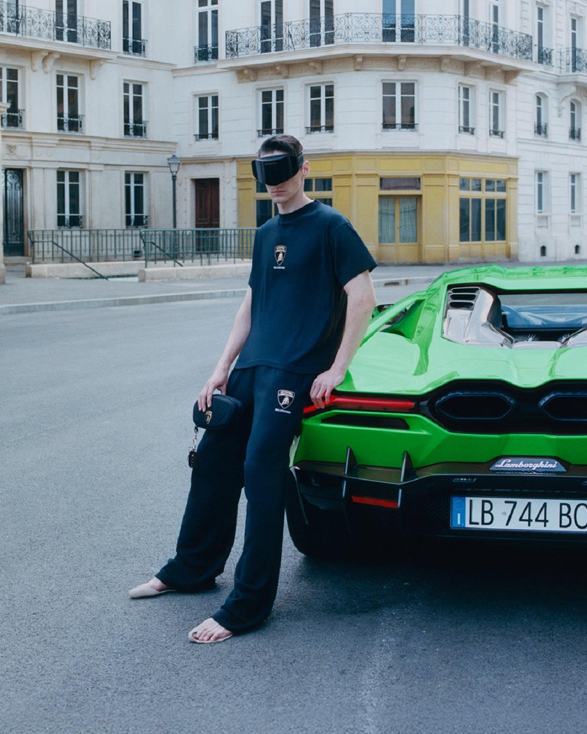 Man dressed in black Lamborghini and Balenciaga branded attire, barefoot, wearing a dark visor, and holding a black Lamborghini pouch, leaning against the rear of a bright green Lamborghini in an elegant urban street setting.