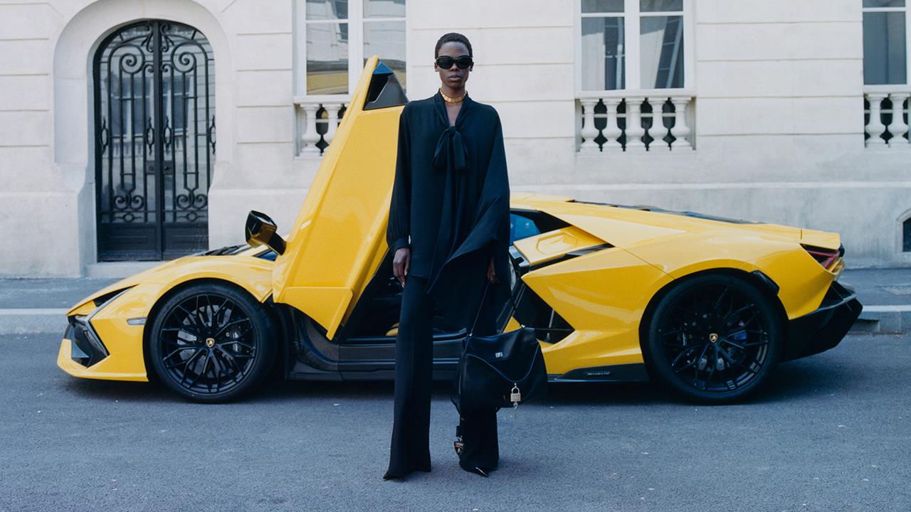 Fashionable individual in black attire standing in front of a bright yellow Lamborghini with scissor doors open, set against a chic urban backdrop.