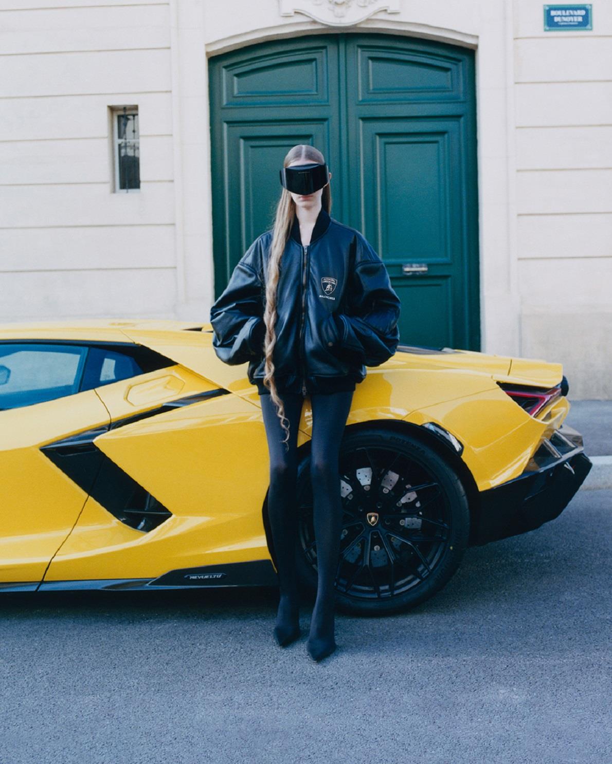 Individual wearing a black Lamborghini-branded jacket, black leggings, pointed shoes, and a futuristic visor, standing in front of a bright yellow Lamborghini Revuelto, set against ornate green double doors and a "Boulevard de l'Université" street sign in an elegant urban setting.