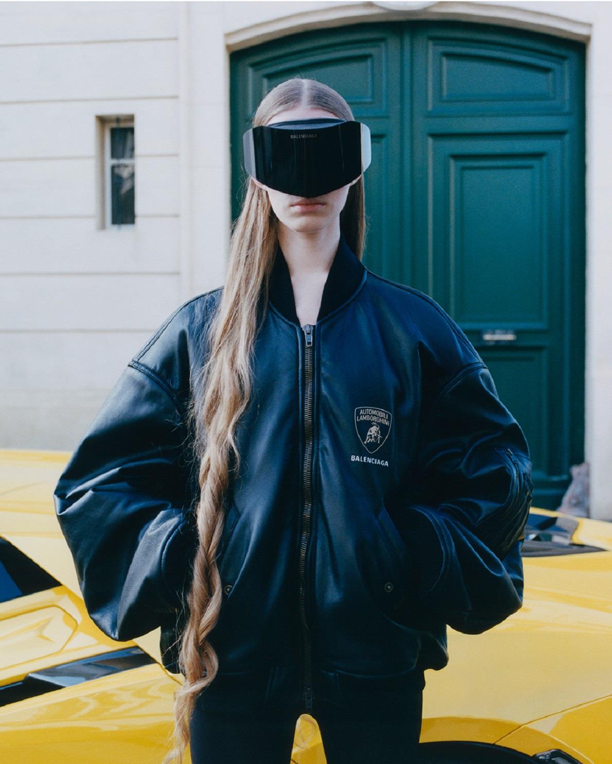 Fashion-forward individual wearing a black leather jacket with Balenciaga and Lamborghini logos, paired with a black visor, standing in front of a green door and a partially visible yellow Lamborghini.