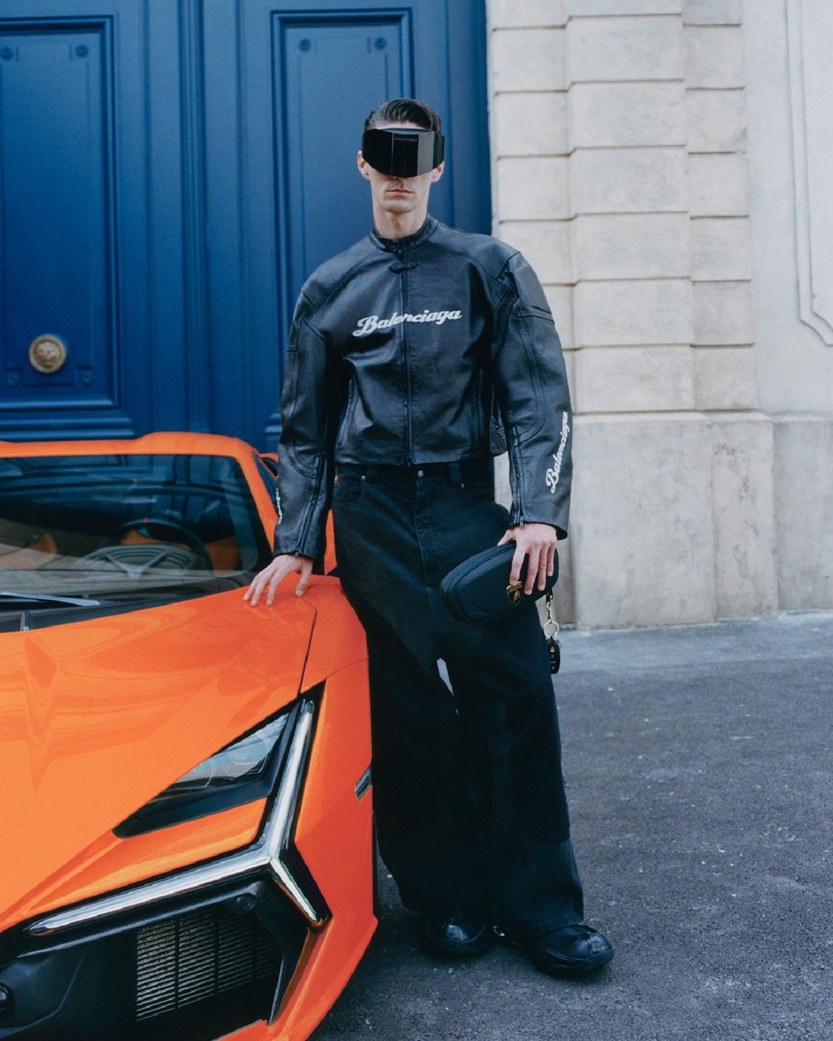 Stylish individual dressed in a black Balenciaga leather jacket with matching pants and futuristic visor, holding a black bag, standing in front of a blue ornate door with an orange luxury sports car partially visible on the side.