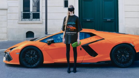 Fashionable individual wearing an oversized black T-shirt with Lamborghini branding, paired with black leggings, pointed shoes, and a black visor, holding a green clutch with a Lamborghini logo, standing in front of a bright orange Lamborghini and a green door in an urban setting.