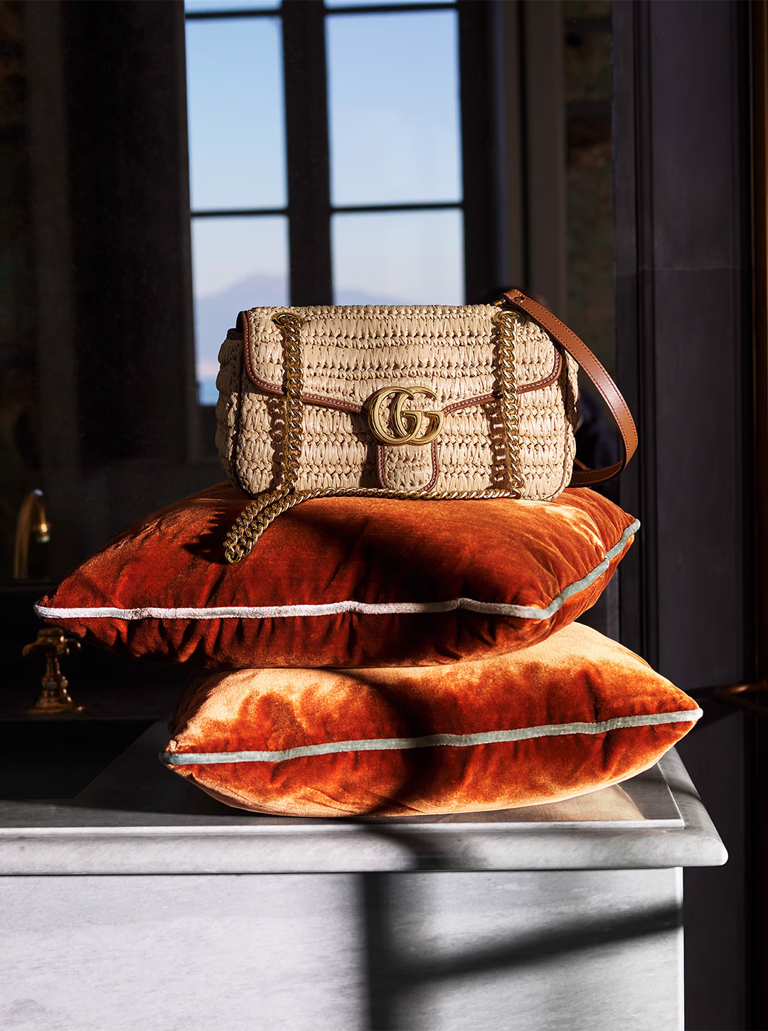 Beige woven Gucci handbag with gold 'GG' logo, chain strap, and dark trim on orange velvet pillows by a window with mountain view.