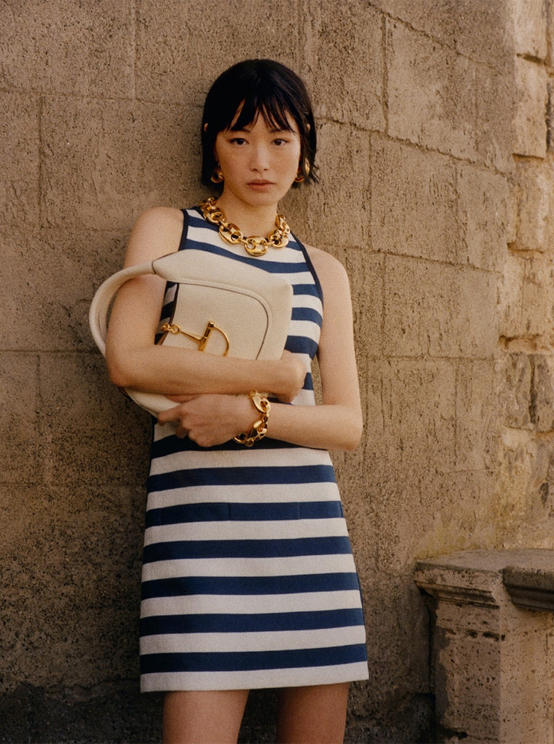 Woman in a Gucci navy and white striped dress with gold jewellery holding a light handbag against a textured stone wall.