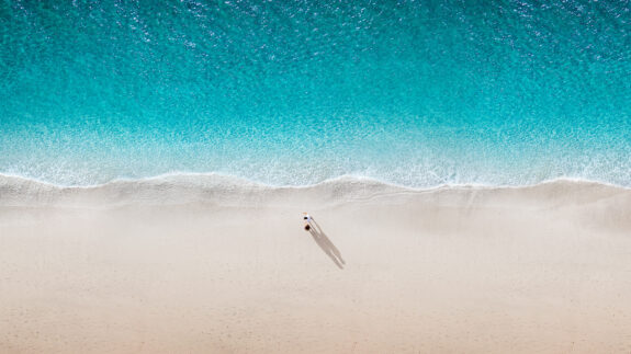 An aerial view of a serene beach with turquoise ocean waves meeting smooth beige sand, where two people walk, casting long shadows in the sunlight.