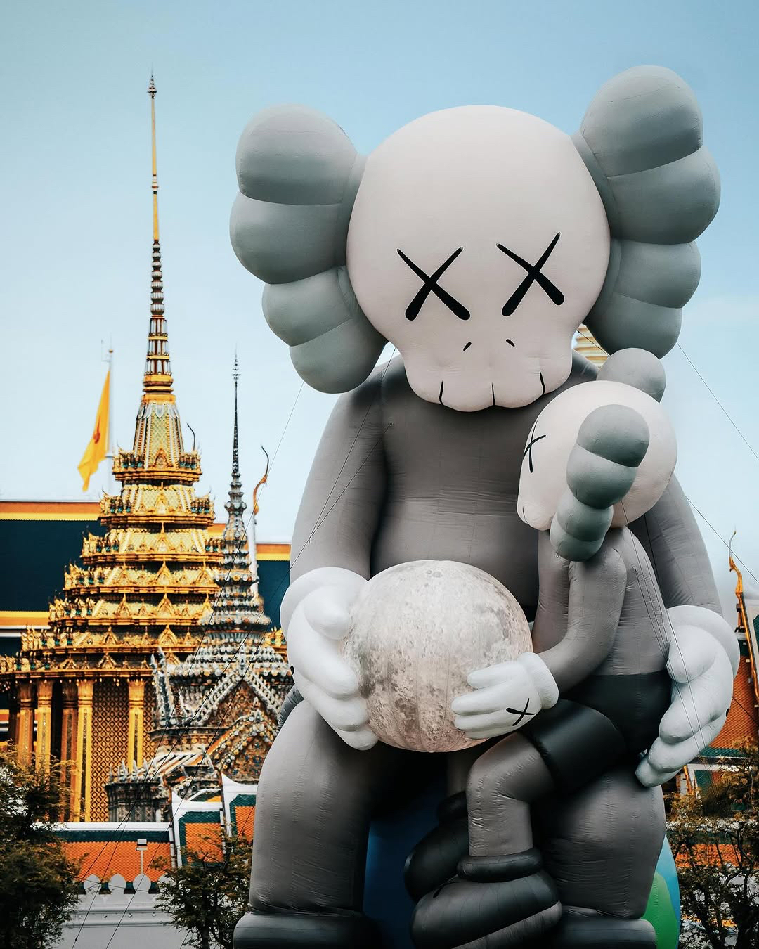 Two large KAWS inflatable figures with X-eyed skull faces, one seated holding a moon-like sphere and the other sitting on its lap, set against the ornate golden spires of a traditional Thai temple.