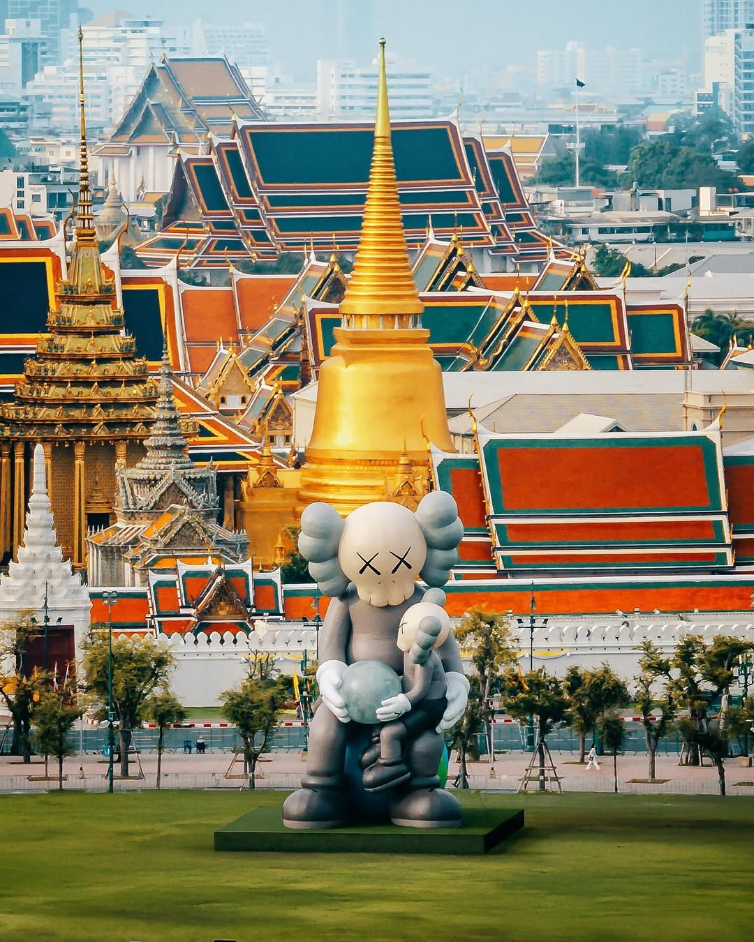 Large KAWS statue with a skull-like face and X-eyed features holding a smaller figure, set against vibrant traditional Thai temple architecture with intricate red, green, and gold designs and a prominent golden spire.