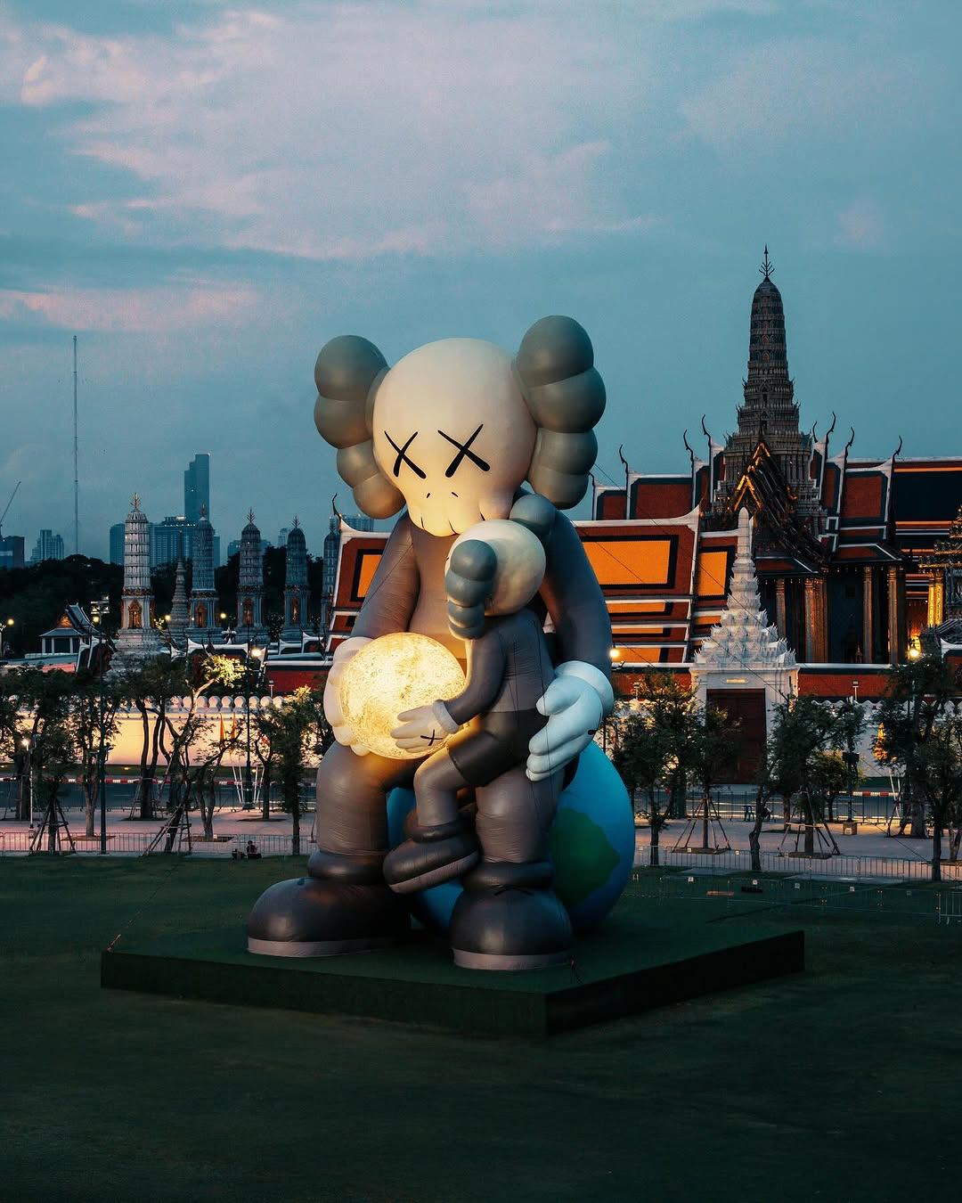 Two large KAWS figures with X-eyed features, one seated holding a smaller figure with a glowing orb, set on a grassy platform against a backdrop of traditional Thai architecture with tiered roofs and intricate designs, with modern skyscrapers visible in the distant background.