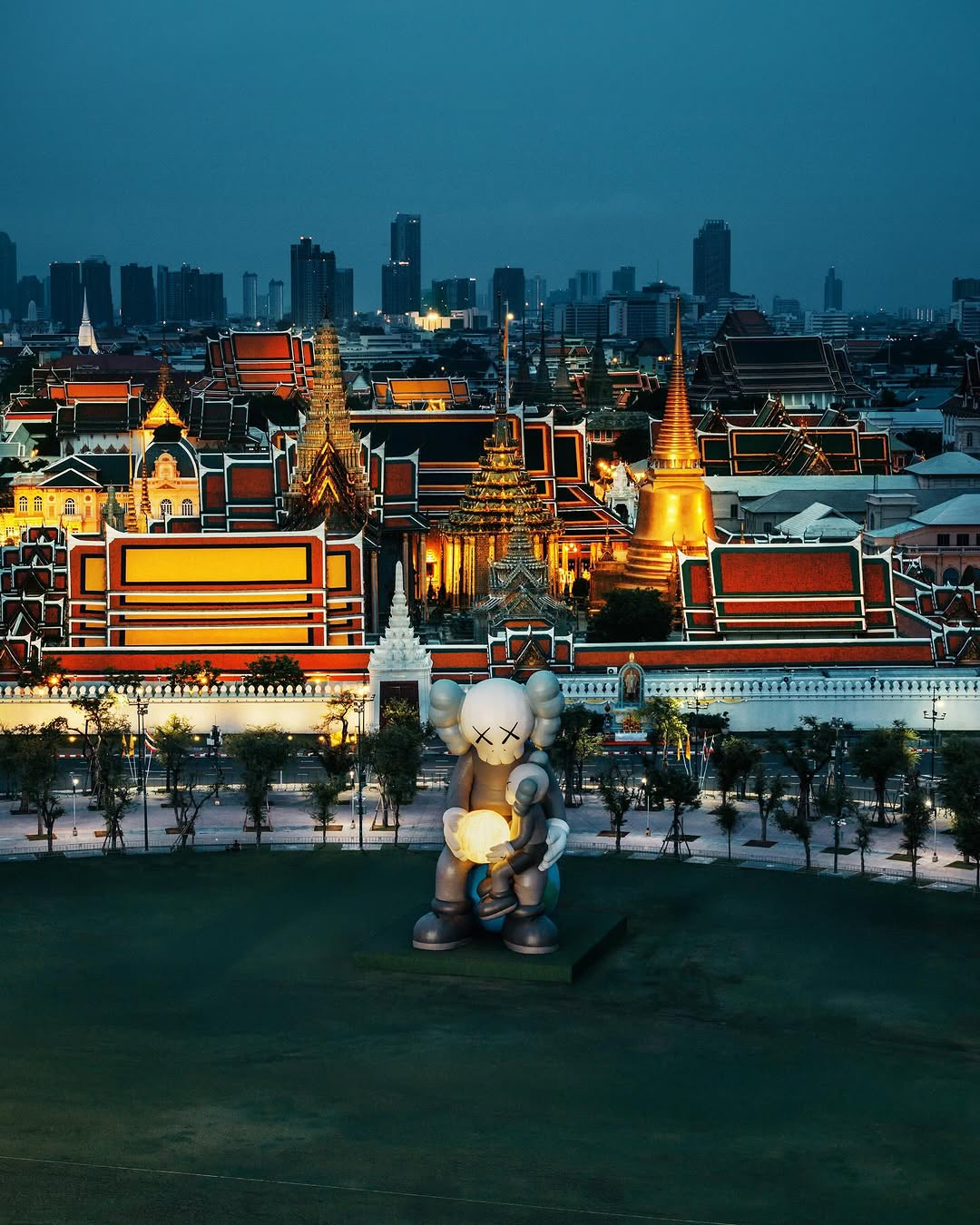 Large KAWS sculpture with X-eyed features holding a glowing orb, set on a grassy area in front of illuminated traditional Thai architecture with tiered roofs and spires, against a backdrop of a modern city skyline at dusk.