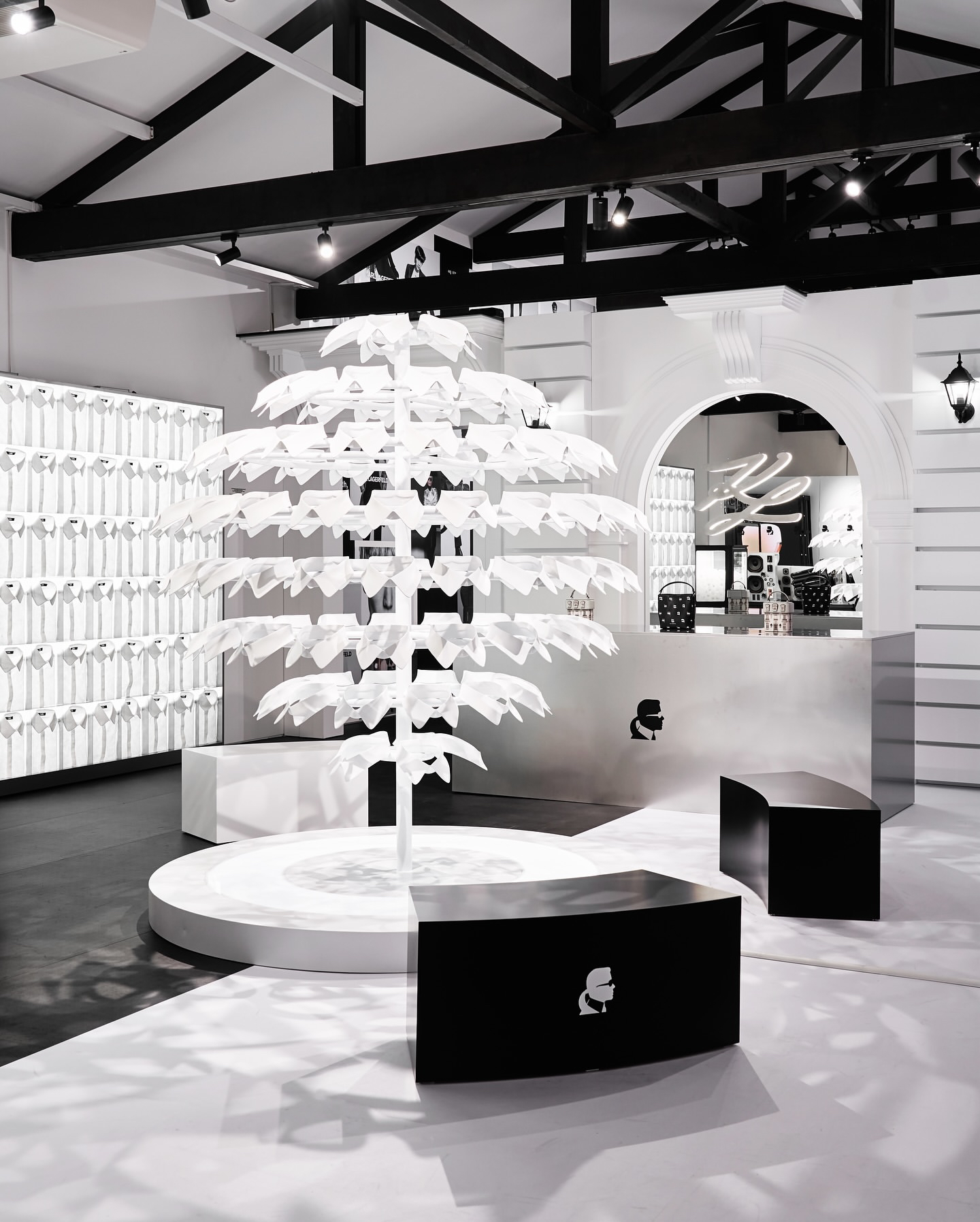 Modern interior of Karl Lagerfeld's immersive Seoul pop-up featuring a central white tree-like structure, black curved benches with a silhouette logo, and monochrome branding elements