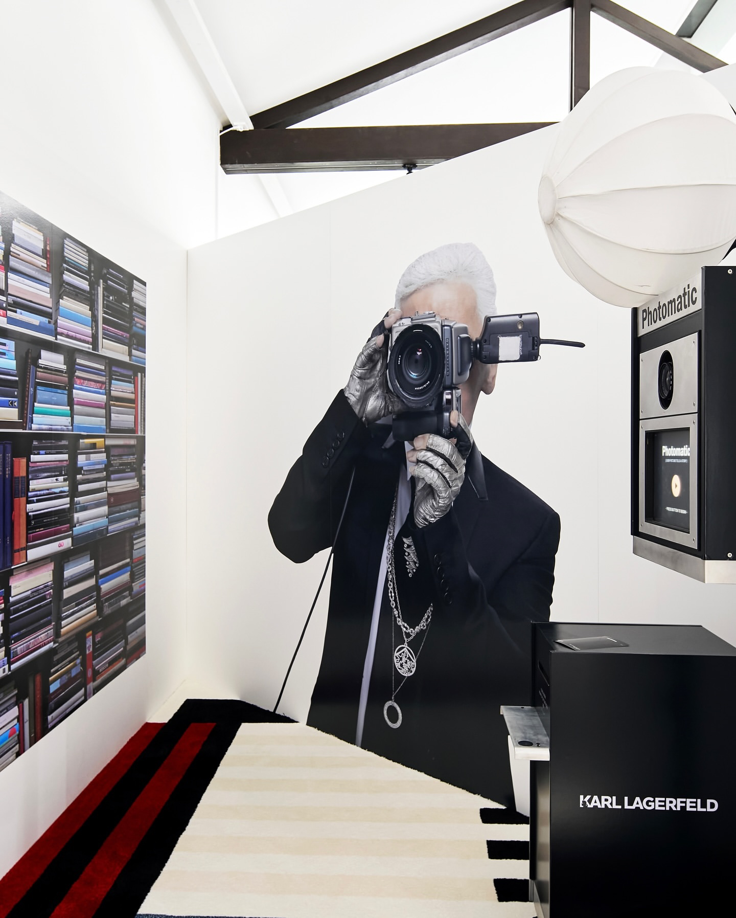 Art installation featuring a large mural of a person with a camera, a Photomatic photo booth, a bookshelf filled with books, and Karl Lagerfeld branding in a modern, elegant setting with striped flooring and creative lighting