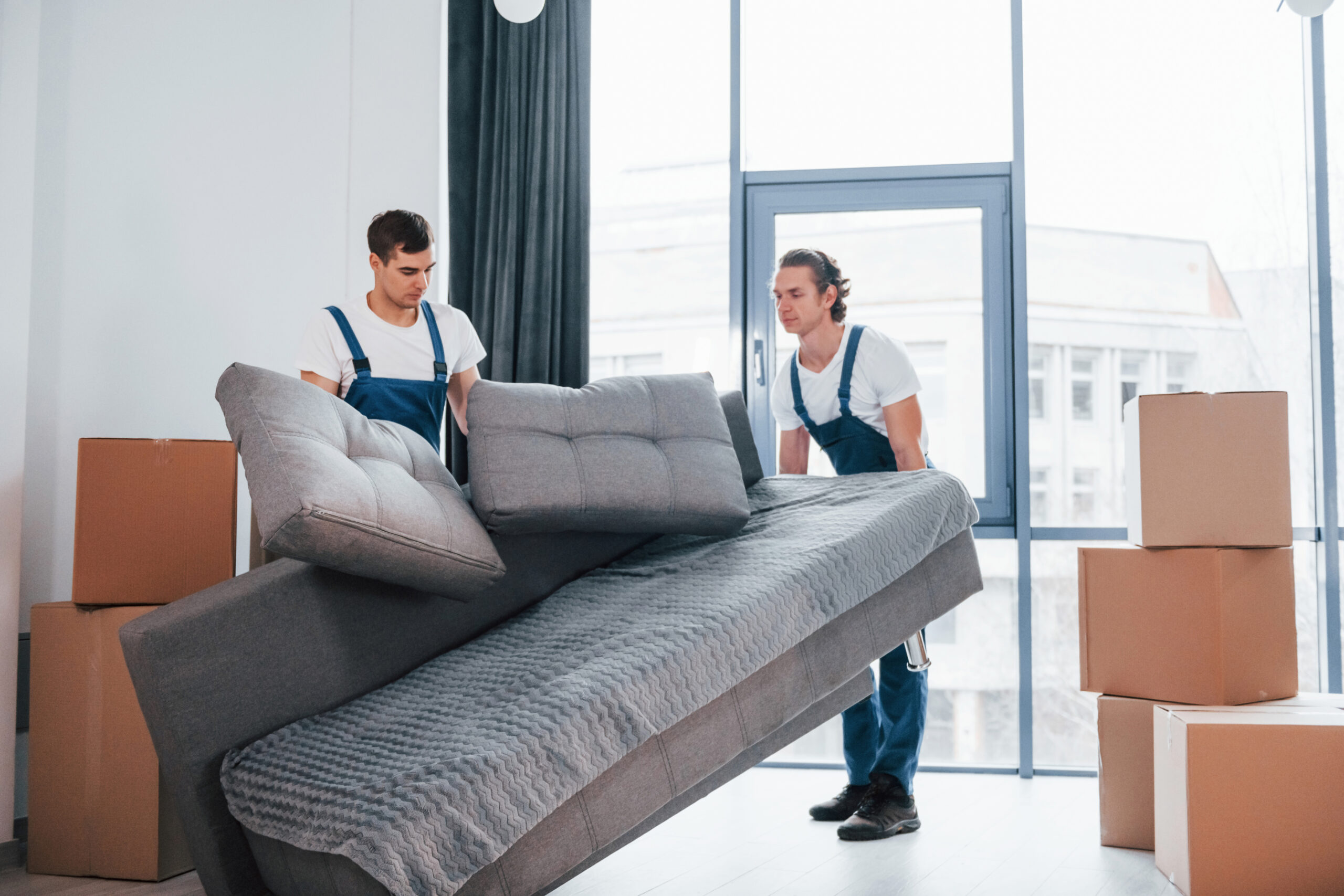 Carrying heavy sofa. Two young movers in blue uniform working indoors in the room.